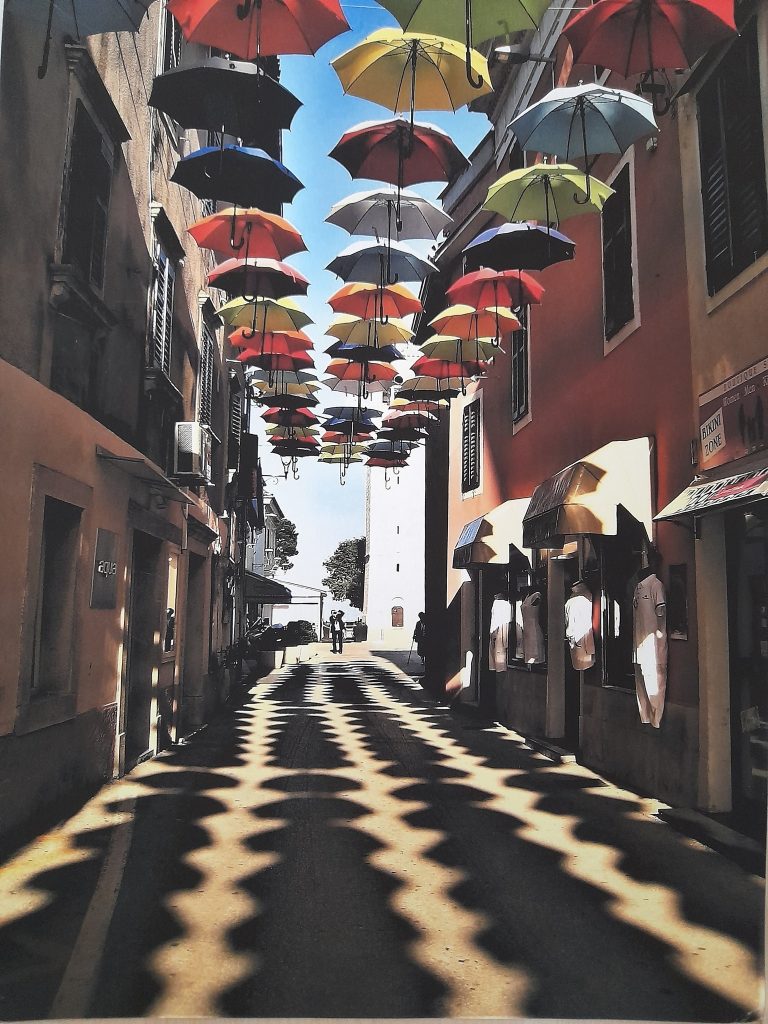 Fotografija uske ulice iznad koje su pričvršćeni kišobrani koji bacaju sjenu na tlo